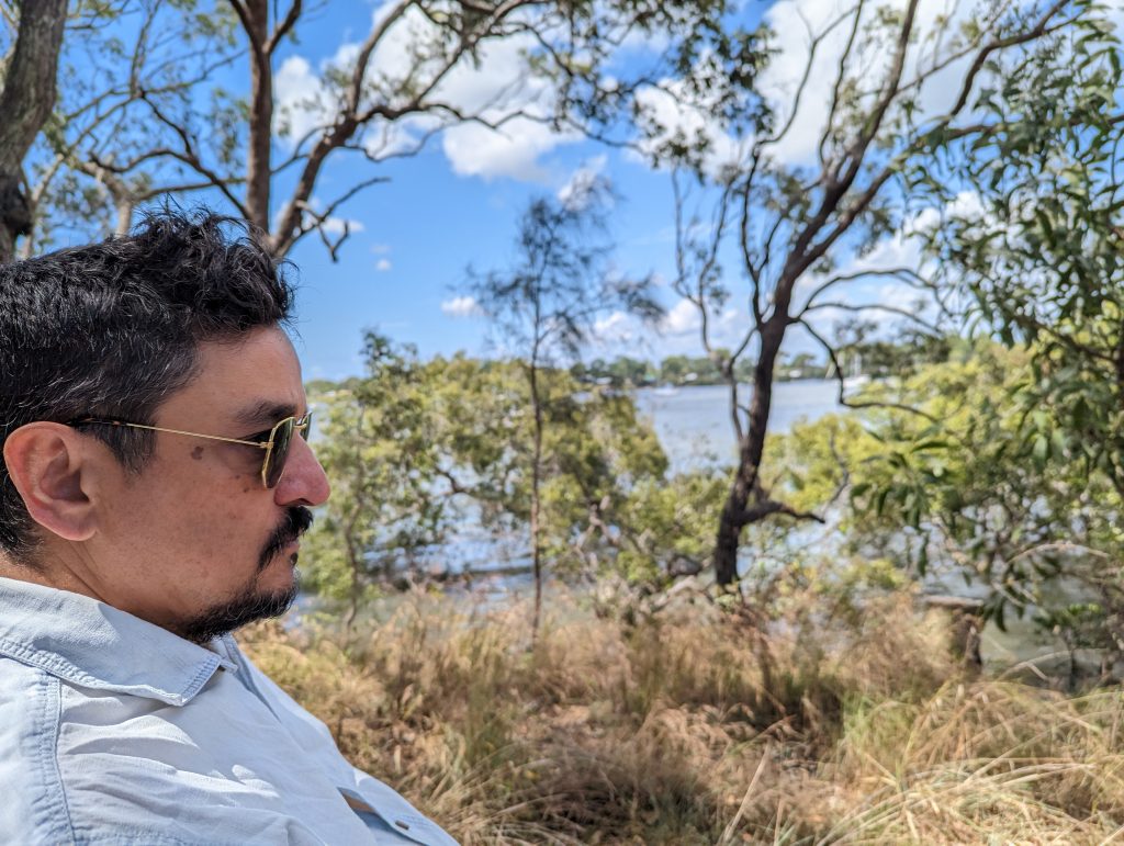 Dev Mukherjee in profile on a sunny day with a background of trees and a bay of water behind them.